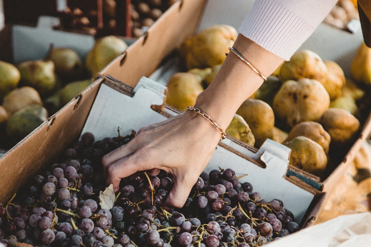 A woman wearing Michael M bracelets grabs a bundle of red grapes at a farmer’s market.