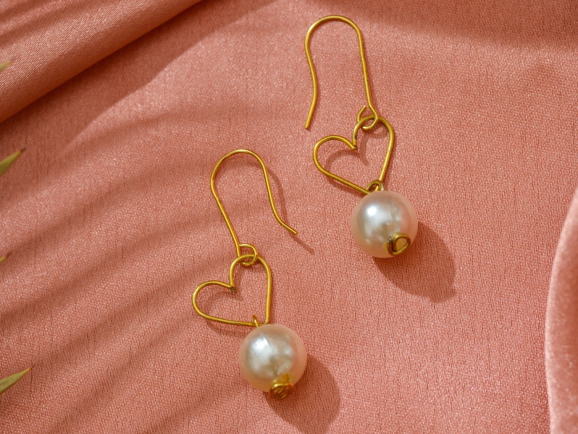 A pair of gold heart drop earrings with pearls on a salmon background