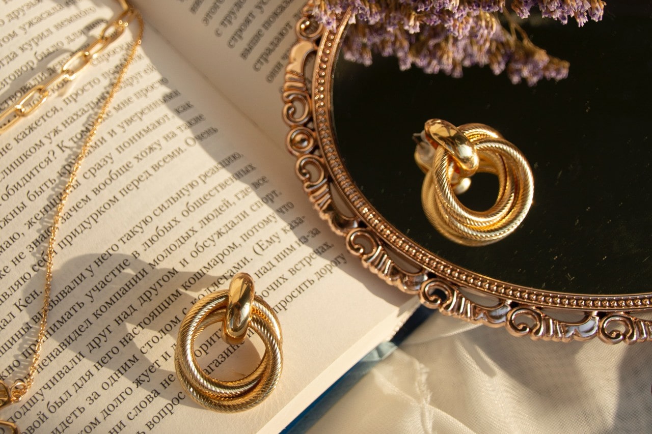 A pair of round gold earrings lying on a mirror and an open book
