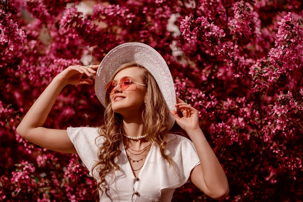 Rose colored glasses, pearls, gold necklaces, and earrings on a woman smiling in front of a flowering tree