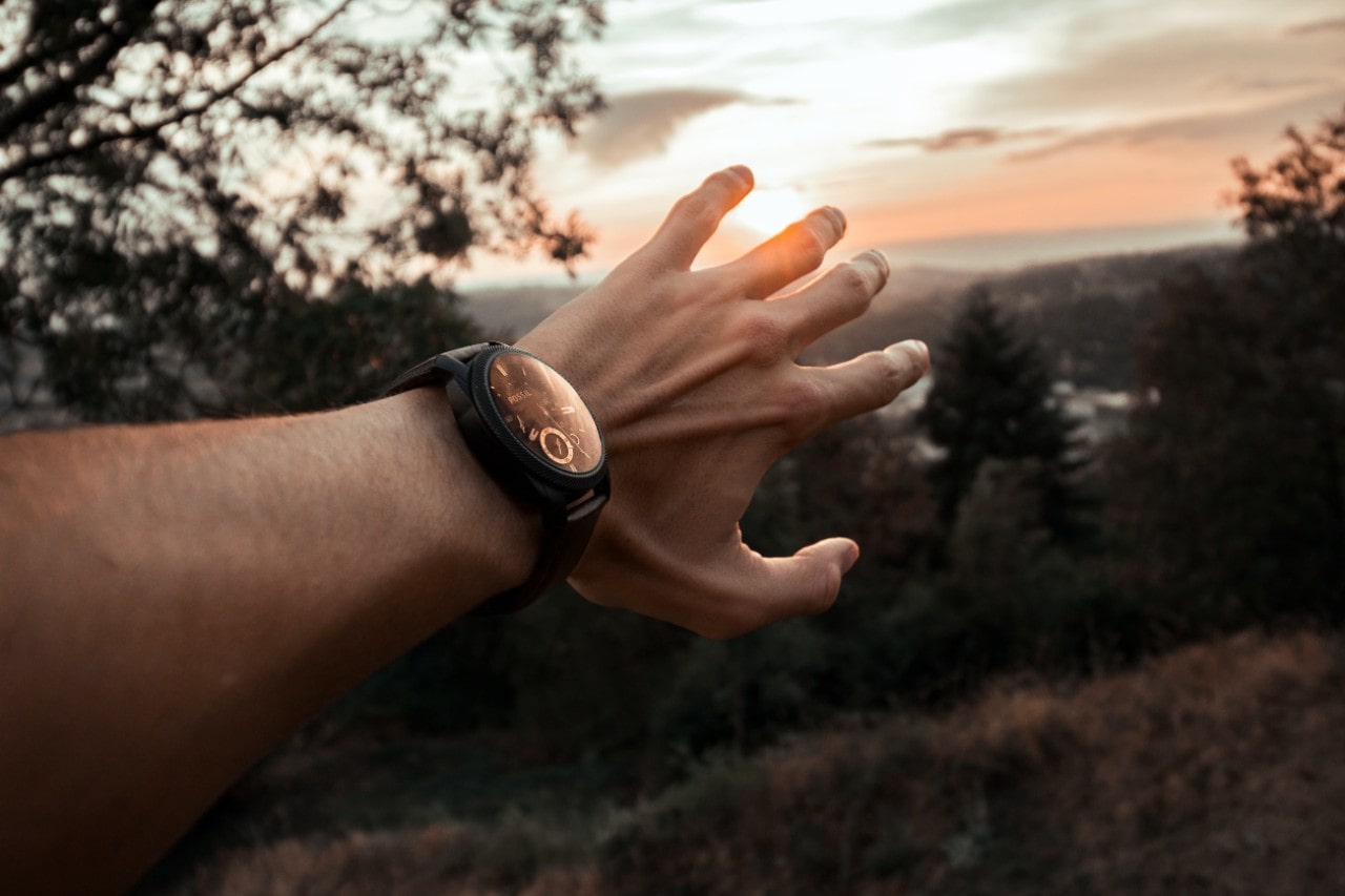 An outstretched arm reaching towards a sunset and wearing a black sports watch