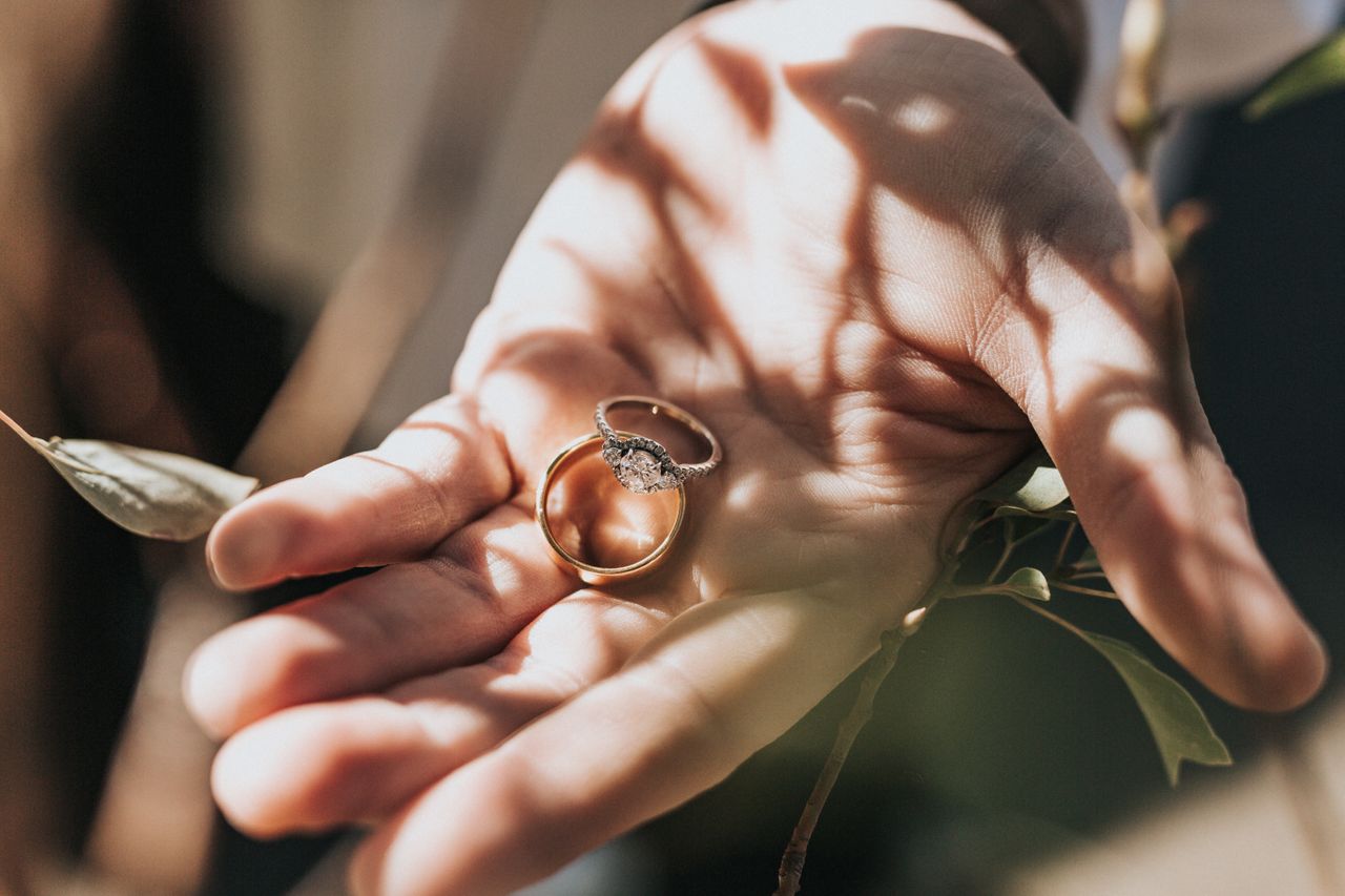 A man holds a yellow gold three-stone engagement ring and a matching wedding band under a tree. A man holds a yellow gold three-stone engagement ring and a matching wedding band under a tree.