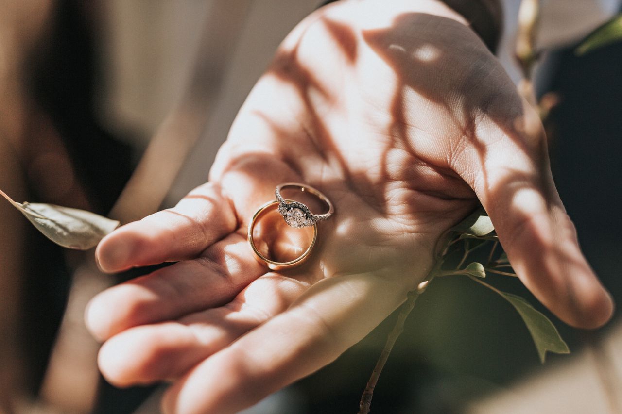 A man holds a yellow gold three-stone engagement ring and a matching wedding band under a tree.