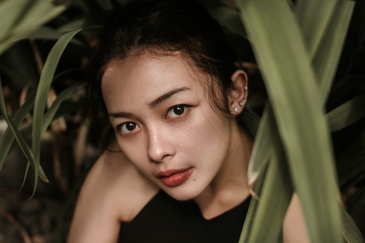 a woman peeking out from behind some plants, wearing a number of diamond stud earrings