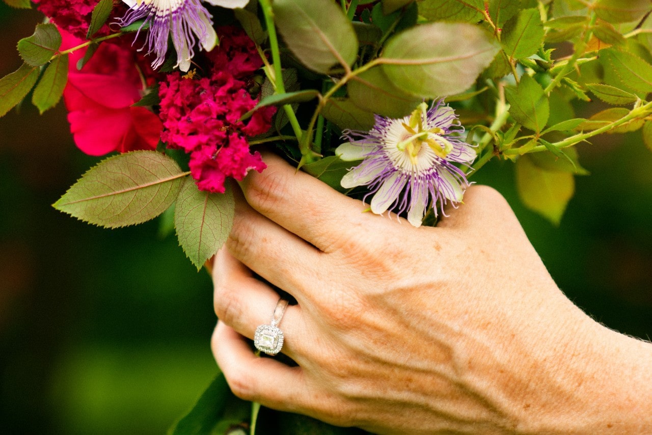 a hand wearing a halo engagement ring and a colorful bouquet of flowers