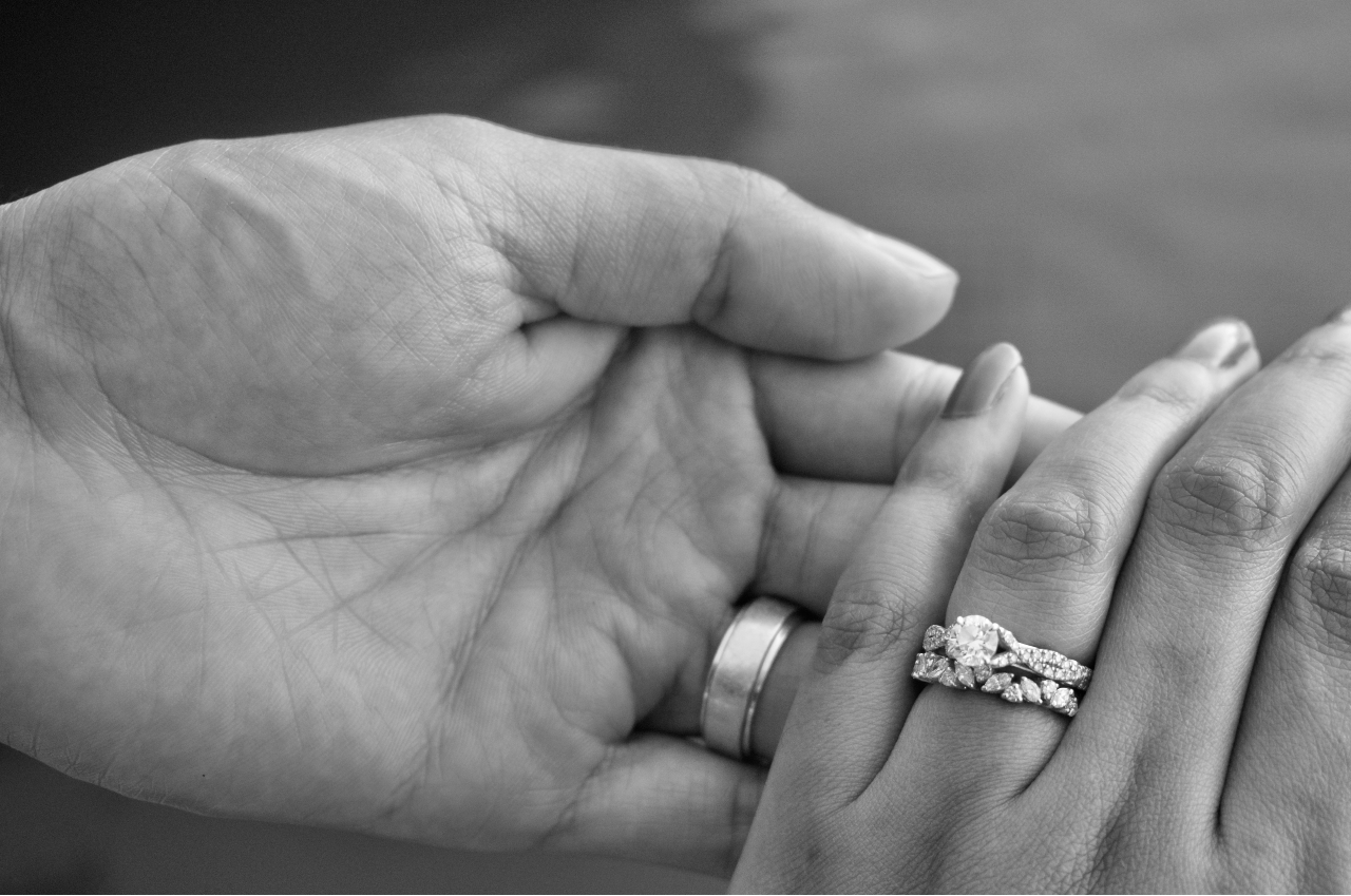 close up image of a man and a woman’s hand, the woman wearing an engagement ring and wedding band