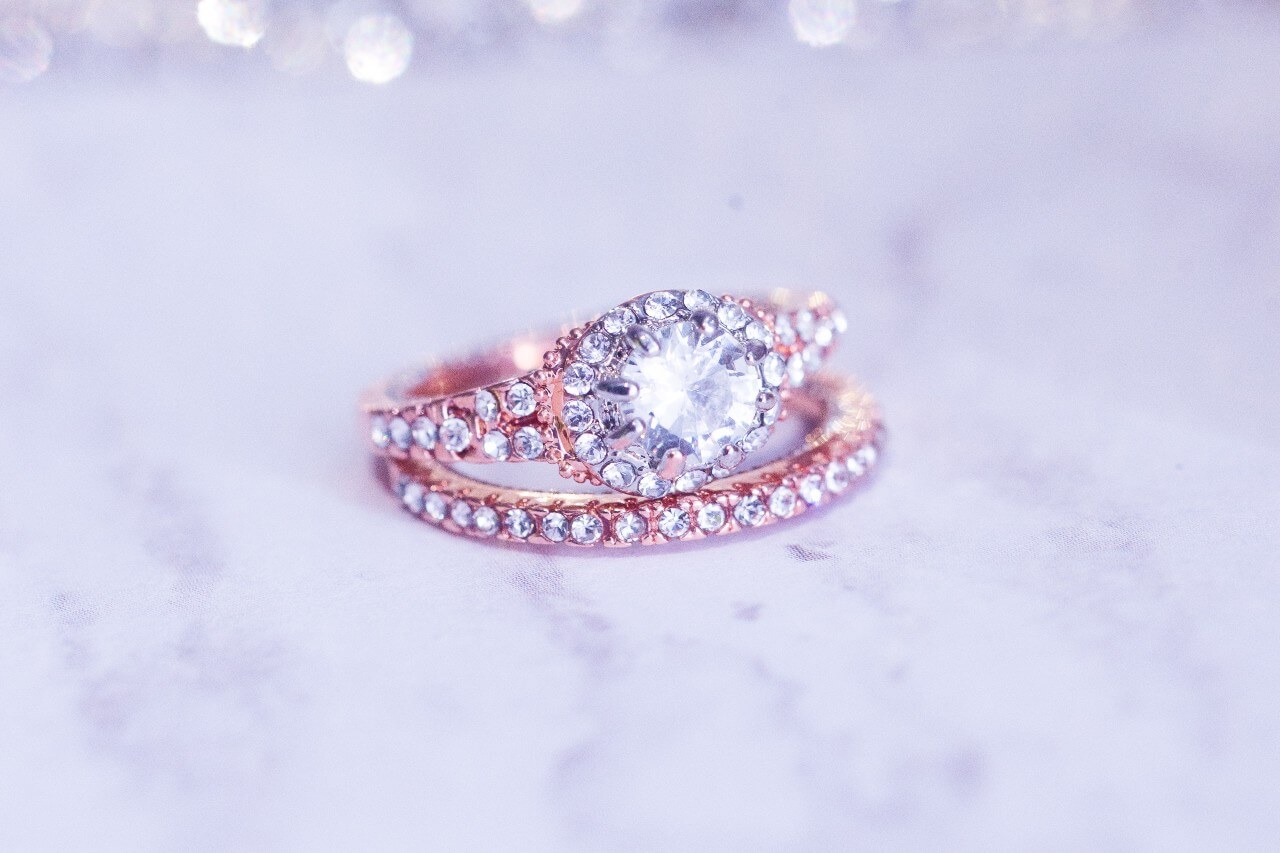 A woman's rose gold wedding ring set with side diamonds and a halo sits on a marble background